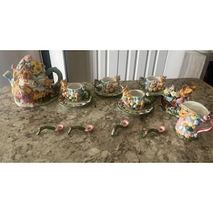 May include: A whimsical ceramic tea set featuring a teapot, teacups, saucers, and spoons. The set is decorated with colorful floral and animal designs. The teapot measures approximately 10 inches in height.