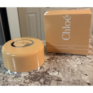 May include: A tan-colored perfume container and box set. The container is round with a silver base and a tan lid with the brand name "Chloé" printed on top. The box is tan with the brand name "Chloé" printed vertically. The box also has the words "perfumed body lotion" printed on it.