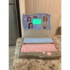 Puede incluir: Un ordenador portátil de juguete electrónico Bratz plateado y rosa. El portátil tiene un teclado rosa, una pantalla con gráficos de Bratz y el texto "POWER ON". El portátil está abierto y colocado sobre una encimera.