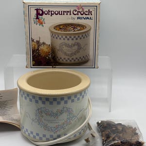 May include: A Rival Potpourri Crock electric warmer with a cream-colored base, a blue and white checkered border, and a floral heart design. Includes the original box with product text, a white power cord, and a small bag of dried potpourri.