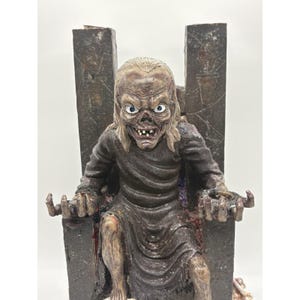 Puede incluir: Una figura detallada de resina de un personaje esquelético de piel pálida con cabello largo, vistiendo una túnica oscura y texturizada. El personaje está sentado en un trono rectangular alto y oscuro con patrones grabados.