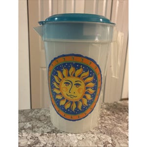 May include: A clear plastic pitcher with a turquoise lid and handle. The pitcher features a colorful sun design with a smiling face on the front. The pitcher is designed for serving beverages and has a capacity of approximately 64 ounces.