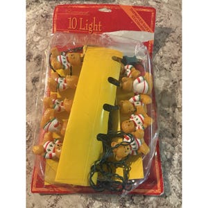 May include: A package of 10 light Christmas string lights. The lights are shaped like small teddy bears wearing red and white sweaters. The package has a red and yellow design with the text "10 Light" and "Improved Safety Standards".
