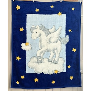 May include: A blue baby blanket featuring a white Pegasus on a cloud within a light blue rectangle. The blanket is adorned with yellow star accents and a dark blue border. The Pegasus has gray wings and mane.