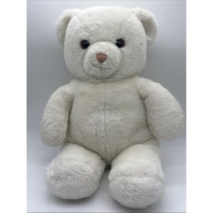 Può includere: Un orsacchiotto di peluche con morbida pelliccia bianca, occhi in plastica marrone scuro e un naso cucito marrone chiaro. L'orso è in posizione seduta con orecchie arrotondate, un muso arrotondato e arti spessi e soffici.