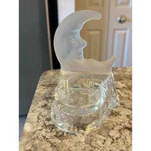 May include: A clear glass candle holder shaped like a crescent moon with a face. The base has a star pattern. The moon is frosted, and the candle holder is clear. The candle holder is on a countertop.