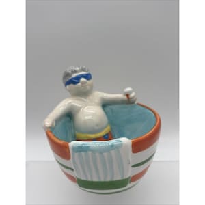 May include: A ceramic bowl featuring a sculpted figure wearing blue sunglasses and yellow swim trunks, holding a small white object. The bowl has a light blue interior and an exterior decorated with horizontal orange and green stripes and a towel.