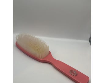 May include: A pink hair brush with a long handle and a rectangular head filled with white bristles. The handle has a rounded end and a silver-colored logo. The brush is designed for detangling and styling hair.