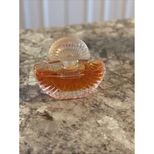 May include: A small, half-circle shaped perfume bottle with a clear, textured glass body and a frosted, mushroom-shaped stopper. The bottle contains a warm amber-colored liquid.