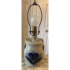 May include: A stoneware lamp with a cream-colored base and a dark blue heart and floral design. The lamp has a metal frame and a light bulb. The lamp is on a speckled countertop.