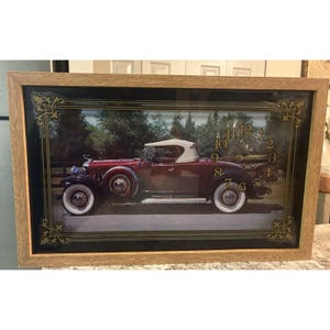 Puede incluir: Un reloj de pared enmarcado con un coche descapotable burdeos vintage con neumáticos blancos y capota blanca. La esfera del reloj tiene números y manecillas doradas. El marco es de madera marrón claro.