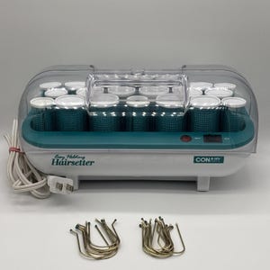 May include: A Conair Easy Holding Hairsetter with a white base, teal interior, and clear plastic lid. The unit contains multiple teal hair rollers and includes two sets of metal U-shaped hair pins. A white power cord is attached to the side.