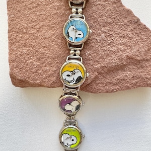 May include: A silver watch with five circular faces, each displaying a different Snoopy design and the text "Circa" followed by a year. The watch is presented on a textured, reddish-brown surface.