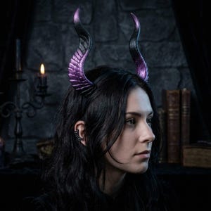 May include: A headband featuring two large, curved horns that transition from a deep black base to a vibrant purple tip. The horns have a textured, ridged surface resembling organic growth, securely attached to a thin, dark, flexible headband frame.