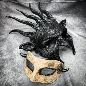 May include: Two ornate masks. The top mask is black with long, curved horns and a textured surface. The bottom mask is tan with black accents and small horns. Both masks have eye openings and are likely for costume or theatrical use.
