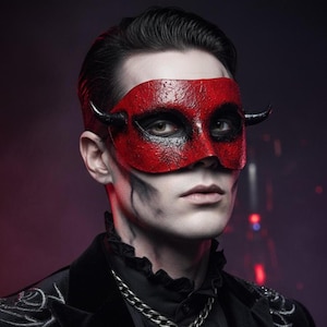 May include: A red and black devil mask with small black horns. The mask covers the eyes and nose, with a textured appearance. The person is wearing a black jacket with a silver chain necklace.