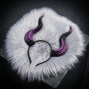 May include: A thin black headband featuring two large, curved horns that transition from a deep black base to a vibrant metallic purple at the tips. The horns have a textured, ridged surface that mimics a natural organic structure with a glossy finish.