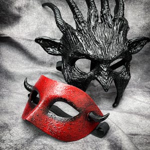 May include: Two theatrical masks. One is a full face mask in black with large horns and a textured surface. The other is a red half-mask with small horns and a textured surface. Both masks have elastic straps.