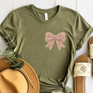 May include: An olive green t-shirt with a pink floral bow applique. The bow has a stitched edge. The shirt has short sleeves and a round neck. A straw hat and sandals are partially visible in the image.