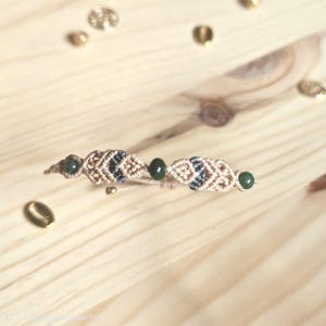 Peut inclure: Un bracelet macramé fait à la main avec un motif tissé beige et bleu marine, orné de perles vert foncé. Le bracelet est présenté sur une surface en bois clair, avec des embellissements dorés éparpillés.