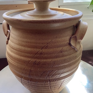 May include: A tan stoneware crock with a matching lid, featuring a textured, ribbed exterior surface with subtle brown speckles and two side handles. The vessel has a wide mouth and a tapered base, showcasing a natural, earthy clay finish.