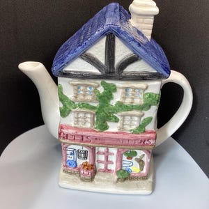 May include: A ceramic teapot shaped like a two-storey building with a blue roof, a white chimney, and green vines climbing the walls. The storefront features a sign reading PETS AND GARDENS, with display windows showing various small items.