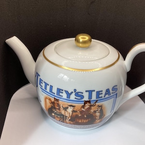 May include: A white ceramic teapot featuring the blue text "TETLEY'S TEAS" above a graphic illustration of four cats playing musical instruments. The teapot has a gold-coloured lid knob and a gold trim around the lid and the upper body.