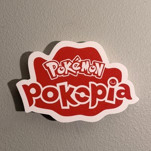 May include: A red and white sticker with the text "Pokémon Poképia" in a stylized font. The sticker has a unique shape, with the text arranged within a red outline. The background is a solid gray color.