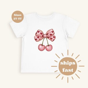Op de afbeelding: Wit T-shirt met een roze strik versierd met rode hartjes en twee roze kersen. Het shirt heeft de tekst "Sizes 2T-5T" en "ships fast" in een cirkelvormig ontwerp.