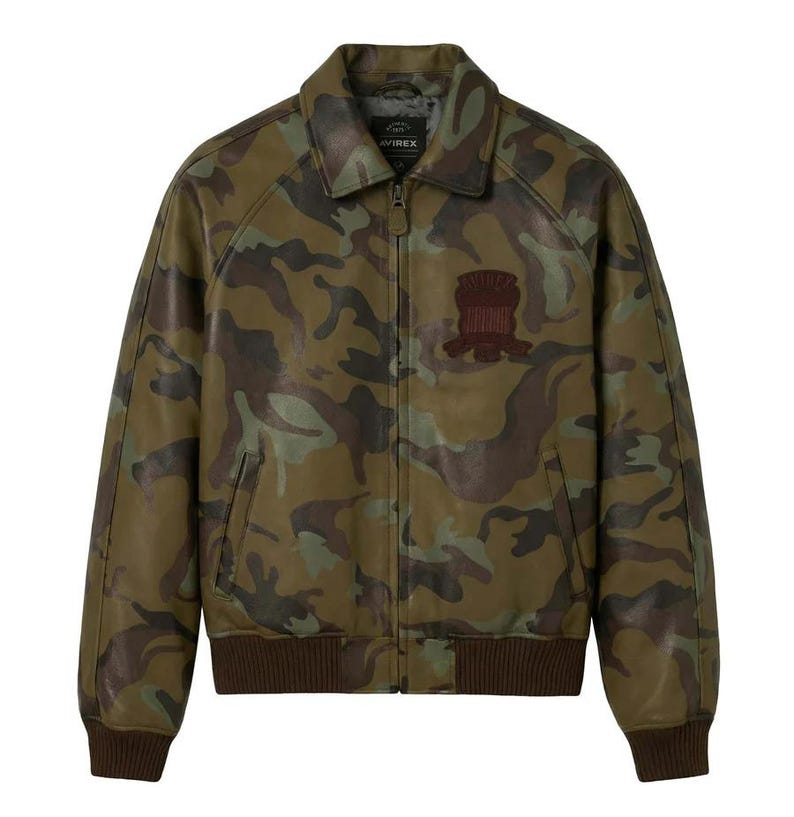 May include: A camouflage-patterned leather jacket in shades of green, brown, and tan. The jacket features a collared neckline, a zipper closure, and a brown ribbed hem and cuffs. A brown patch is sewn on the chest. The brand name is visible on the collar.