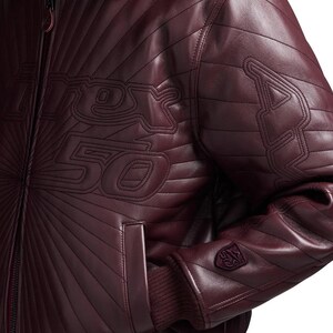 May include: Burgundy leather jacket with quilted detailing and the text "Rex 50" stitched across the chest. Features a ribbed cuff and a small emblem on the sleeve. The jacket has a zip-up front and a classic bomber style.