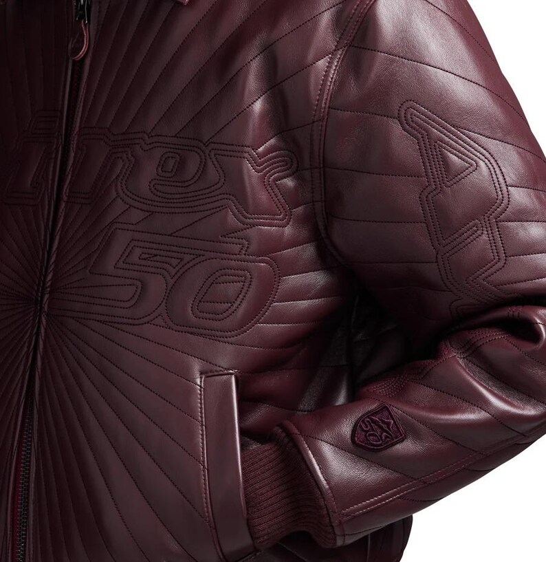 May include: Burgundy leather jacket with quilted detailing and the text "Rex 50" stitched across the chest. Features a ribbed cuff and a small emblem on the sleeve. The jacket has a zip-up front and a classic bomber style.