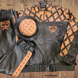 May include: A black and tan leather set featuring a zip-up jacket, matching joggers, and a bucket hat. The jacket and joggers display the MCM logo and a diamond pattern. The bucket hat has a tan crown with the MCM logo and a black brim.
