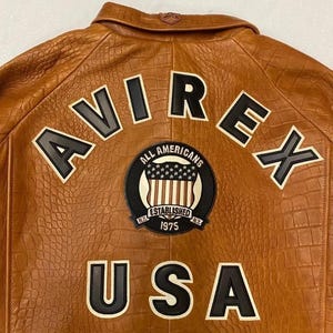 May include: Brown leather jacket with embossed texture. Features the word "AVIREX" across the back in black and white lettering, along with a circular patch that reads "ALL AMERICANS ESTABLISHED 1975 N.Y." and the word "USA".