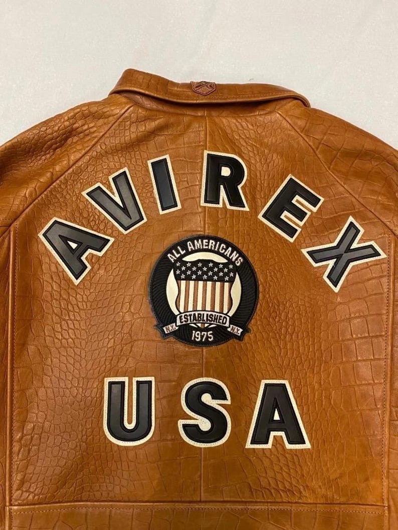 May include: Brown leather jacket with embossed texture. Features the word "AVIREX" across the back in black and white lettering, along with a circular patch that reads "ALL AMERICANS ESTABLISHED 1975 N.Y." and the word "USA".