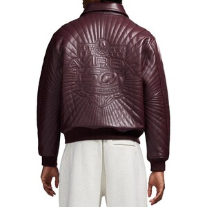 May include: Burgundy leather bomber jacket with a quilted back panel. The back features an embossed design with the text "AVIREX 50 GOLD KICKOFF NY". The jacket has a ribbed collar, cuffs, and hem.
