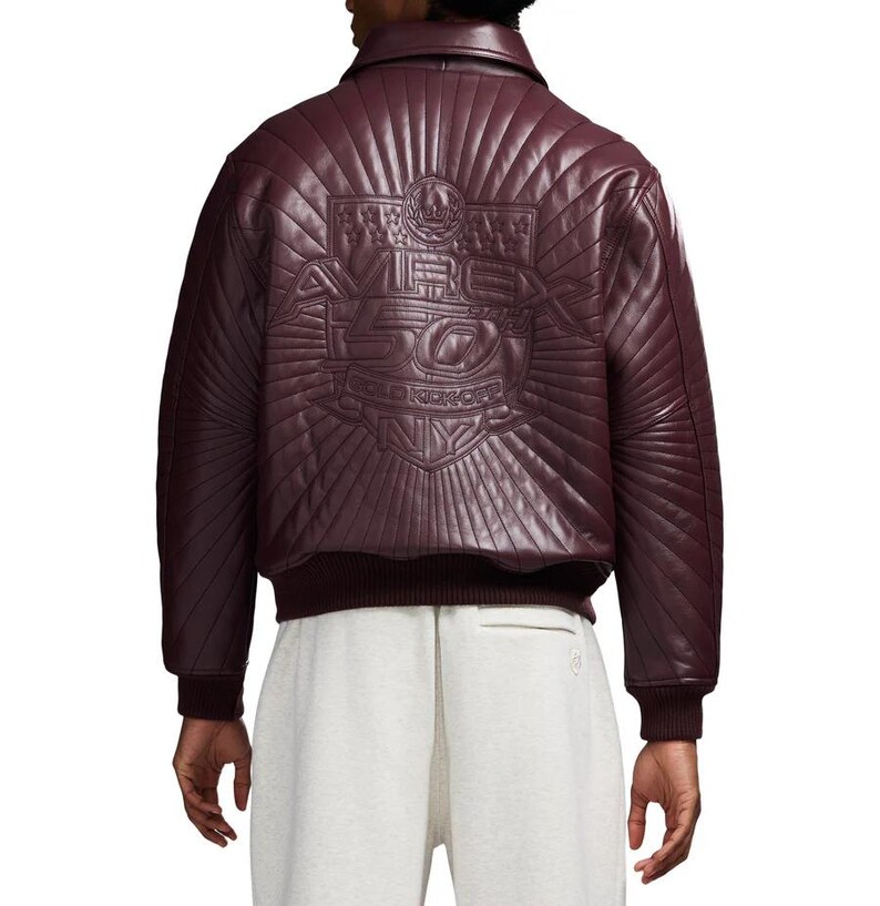 May include: Burgundy leather bomber jacket with a quilted back panel. The back features an embossed design with the text "AVIREX 50 GOLD KICKOFF NY". The jacket has a ribbed collar, cuffs, and hem.