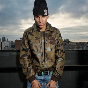 May include: A camouflage-patterned jacket with a brown collar and cuffs. The jacket features a zipper closure and a patch on the chest. The person is wearing a black beanie with a small patch and blue jeans.