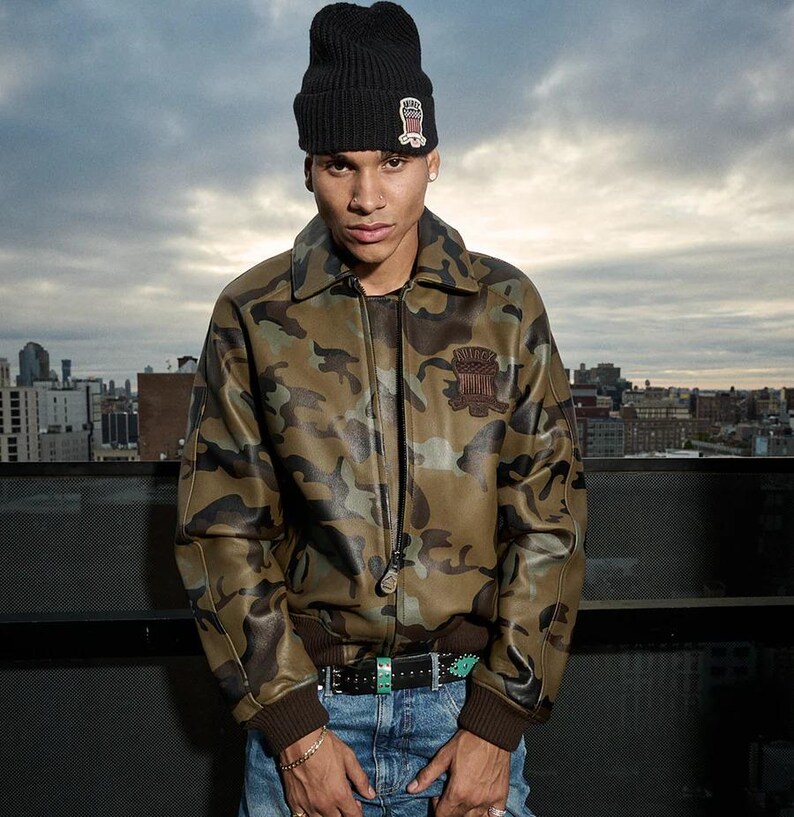 May include: A camouflage-patterned jacket with a brown collar and cuffs. The jacket features a zipper closure and a patch on the chest. The person is wearing a black beanie with a small patch and blue jeans.