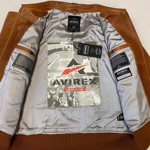 May include: A brown leather jacket with a ribbed hem and cuffs. The interior is lined with a silver fabric and features a graphic print with the text "AVIREX LIMITED EDITION". The jacket has multiple pockets and a zipper closure.
