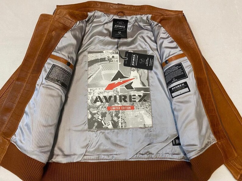 May include: A brown leather jacket with a ribbed hem and cuffs. The interior is lined with a silver fabric and features a graphic print with the text "AVIREX LIMITED EDITION". The jacket has multiple pockets and a zipper closure.