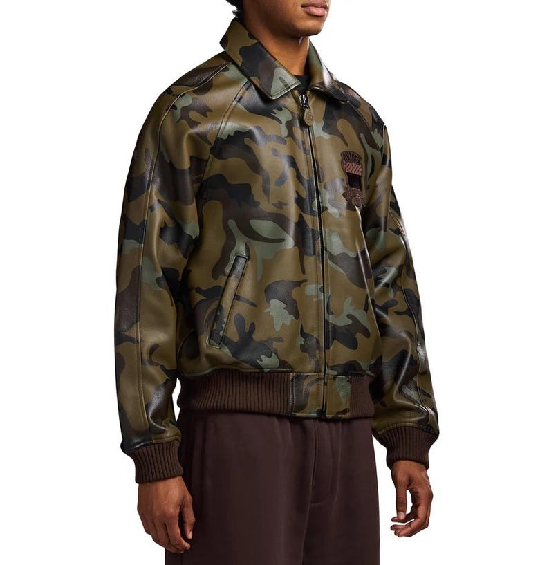 May include: A camouflage-patterned jacket in shades of green, brown, and black. The jacket features a collared neckline, a front zipper, and a brown ribbed hem and cuffs. A small patch is visible on the chest. The jacket appears to be made of a leather-like material.