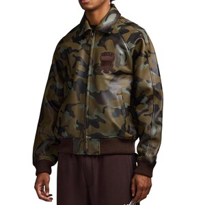 May include: Camouflage-patterned jacket in shades of green, brown, and black. The jacket features a brown collar, cuffs, and waistband. A patch with text is visible on the chest. The jacket has a zipper closure.
