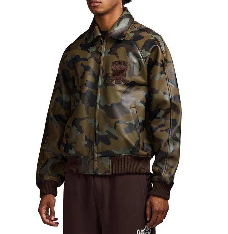 May include: Camouflage-patterned jacket in shades of green, brown, and black. The jacket features a brown collar, cuffs, and waistband. A patch with text is visible on the chest. The jacket has a zipper closure.