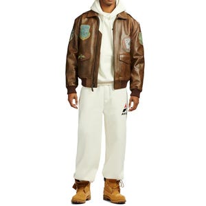Pu&ograve; includere: Giacca bomber in pelle marrone con toppe, indossata sopra una felpa con cappuccio e pantaloni della tuta color crema. La giacca ha una chiusura con zip, tasche e polsini a coste. I pantaloni della tuta hanno un logo sulla gamba sinistra. L'outfit &egrave; completato da stivali da lavoro color cuoio.