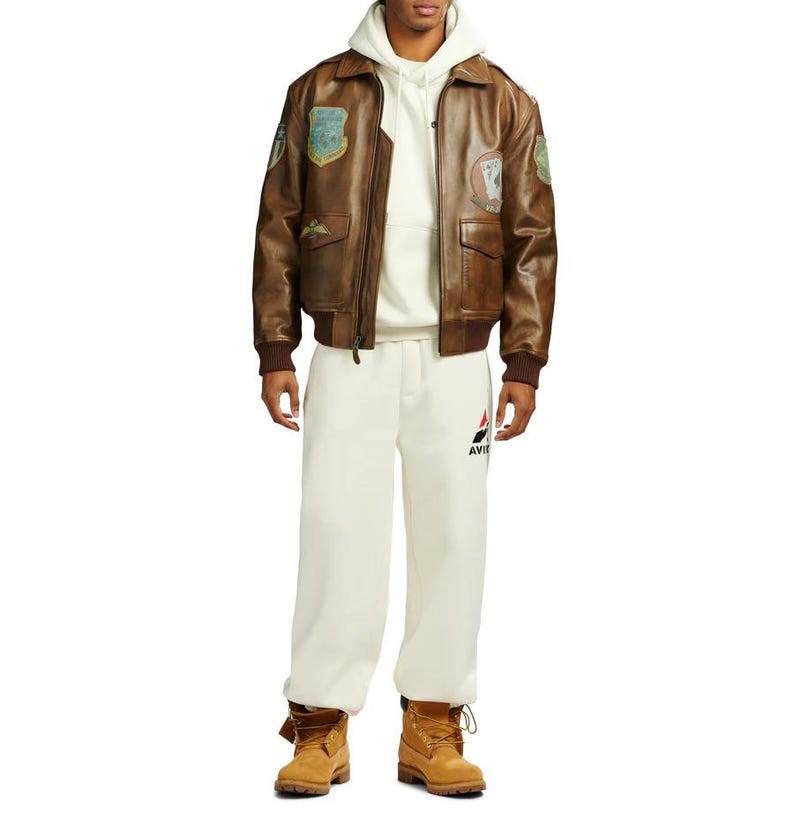 Pu&ograve; includere: Giacca bomber in pelle marrone con toppe, indossata sopra una felpa con cappuccio e pantaloni della tuta color crema. La giacca ha una chiusura con zip, tasche e polsini a coste. I pantaloni della tuta hanno un logo sulla gamba sinistra. L'outfit &egrave; completato da stivali da lavoro color cuoio.