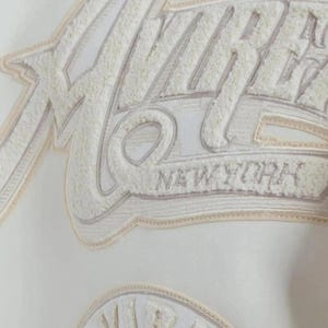 Puede incluir: Primer plano de una tela blanca con un dise&ntilde;o bordado. El dise&ntilde;o presenta la palabra "VIBE" en una fuente texturizada blanquecina, con "NEW YORK" debajo. El bordado tiene un efecto tridimensional en relieve.