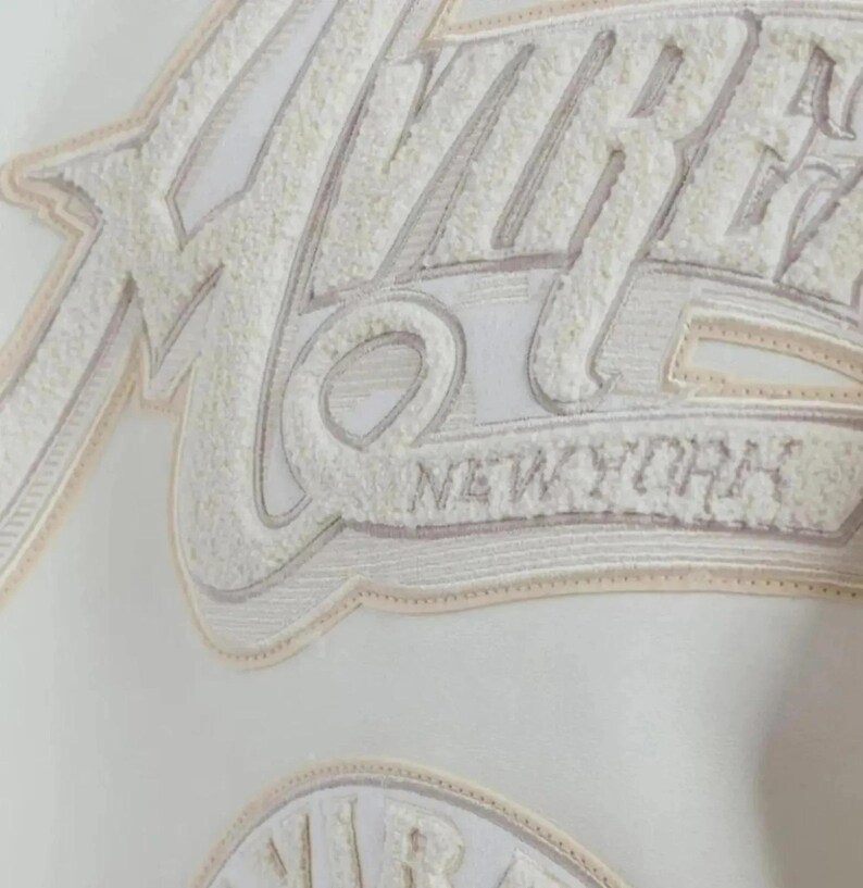 Puede incluir: Primer plano de una tela blanca con un dise&ntilde;o bordado. El dise&ntilde;o presenta la palabra "VIBE" en una fuente texturizada blanquecina, con "NEW YORK" debajo. El bordado tiene un efecto tridimensional en relieve.