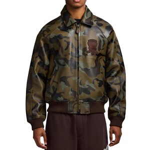 May include: Camouflage-patterned leather jacket in shades of green, brown, and black. The jacket features a collared neckline, a front zipper, and a brown ribbed hem and cuffs. A brown patch is sewn on the upper left chest.