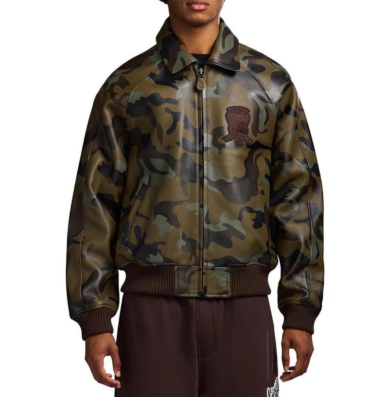 May include: Camouflage-patterned leather jacket in shades of green, brown, and black. The jacket features a collared neckline, a front zipper, and a brown ribbed hem and cuffs. A brown patch is sewn on the upper left chest.
